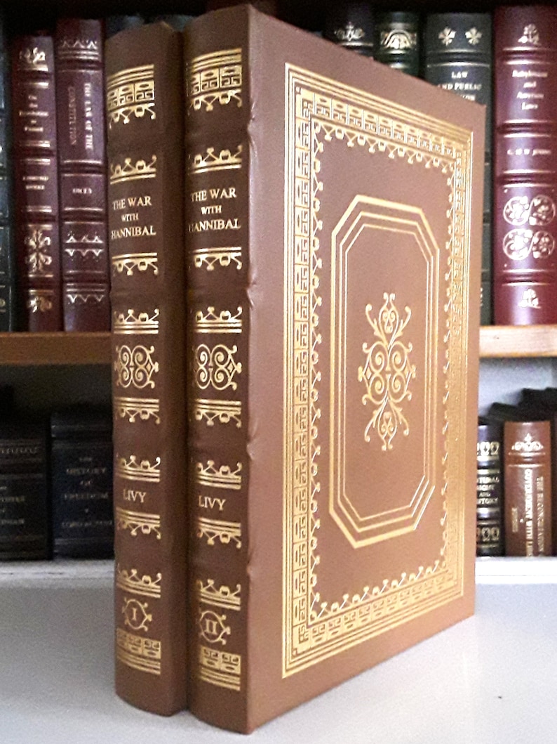 Puede incluir: Dos libros encuadernados en cuero marr&oacute;n con detalles dorados y el t&iacute;tulo "La Guerra con An&iacute;bal" en el lomo. Los libros son de Tito Livio y son los vol&uacute;menes I y II.