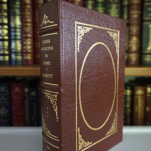 Puede incluir: Un libro encuadernado en cuero marr&oacute;n con detalles dorados y el t&iacute;tulo "Greek Medicine in Rome" de T.C. Allbutt. El libro forma parte de "The Classics of Medicine Library".