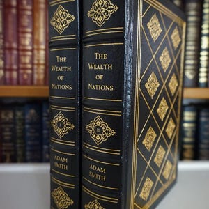 May include: Two hardcover books titled "The Wealth of Nations" by Adam Smith. The books are black with gold decorative accents and text. The spines feature gold filigree designs and the Roman numerals I and II.