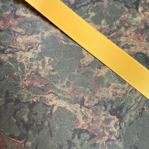 May include: A close-up of a yellow satin ribbon on a green, brown, and pink marbled background.