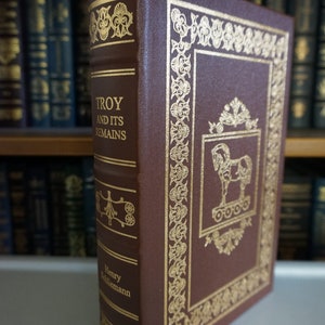 Puede incluir: Un libro encuadernado en cuero marr&oacute;n con letras doradas y detalles decorativos. El t&iacute;tulo es "Troy and Its Remains" de Henry Schliemann.