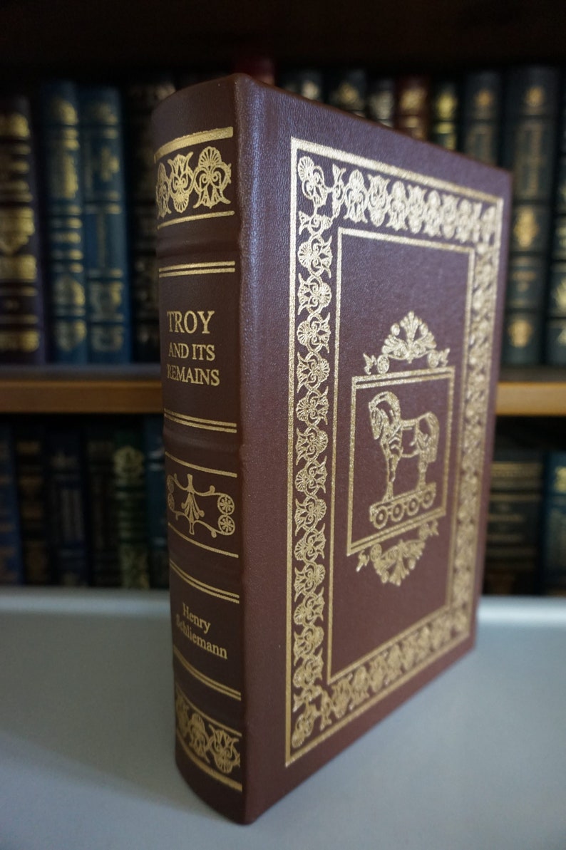 Puede incluir: Un libro encuadernado en cuero marr&oacute;n con letras doradas y detalles decorativos. El t&iacute;tulo es "Troy and Its Remains" de Henry Schliemann.