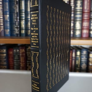 May include: A hardcover book titled "Pride Before the Fall" by John Heilemann. The book has a black cover with gold lettering and decorative gold lines. The book is standing upright in front of a bookshelf.