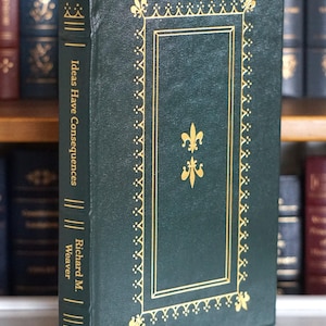 Puede incluir: Un libro de tapa dura verde oscuro titulado "Ideas Have Consequences" de Richard M. Weaver. El libro presenta elementos decorativos dorados, incluyendo una flor de lis y un marco rectangular. Se ve una cinta dorada como marcapáginas.