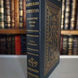 Puede incluir: Un libro de tapa dura de color azul oscuro con letras doradas y detalles decorativos. El t&iacute;tulo es "Agust&iacute;n y los jud&iacute;os" de Paula Fredriksen.