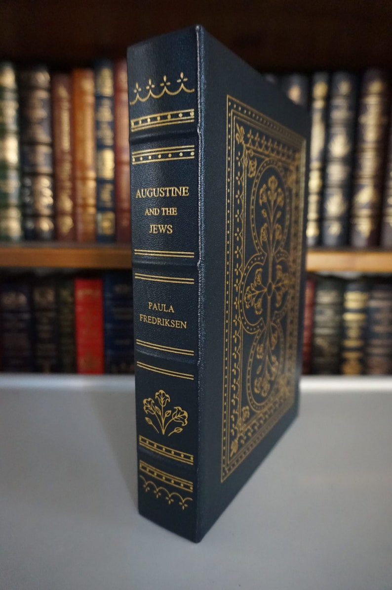 Puede incluir: Un libro de tapa dura de color azul oscuro con letras doradas y detalles decorativos. El t&iacute;tulo es "Agust&iacute;n y los jud&iacute;os" de Paula Fredriksen.