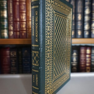 May include: A hardcover book titled "On the Donation of Constantine" by Lorenzo Valla. The book has a dark green cover with gold decorative patterns and lettering. A yellow ribbon bookmark is visible. The background shows a bookshelf filled with other books.