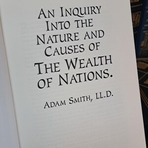 K&ouml;nnte beinhalten: Aufgeschlagenes Buch mit dem Titel "An Inquiry Into the Nature and Causes of the Wealth of Nations" von Adam Smith. Der Text ist in schwarzer Schrift. Im Hintergrund sind weitere B&uuml;cher mit dunklen Einb&auml;nden zu sehen.