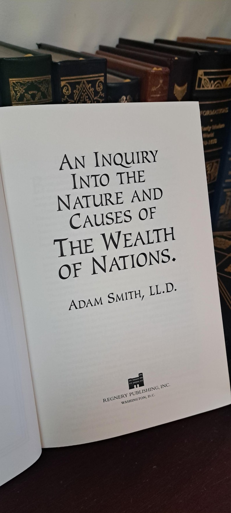 K&ouml;nnte beinhalten: Aufgeschlagenes Buch mit dem Titel "An Inquiry Into the Nature and Causes of the Wealth of Nations" von Adam Smith. Der Text ist in schwarzer Schrift. Im Hintergrund sind weitere B&uuml;cher mit dunklen Einb&auml;nden zu sehen.