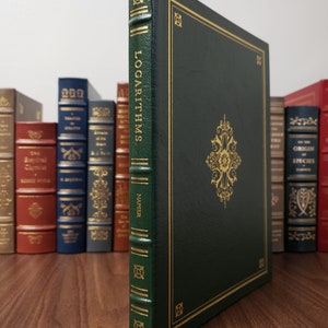 Puede incluir: Un libro verde oscuro titulado "LOGARITHMS" de Napier, con letras doradas y detalles decorativos. El libro tiene un borde dorado y un emblema dorado detallado en la portada. Otros libros son visibles en el fondo.