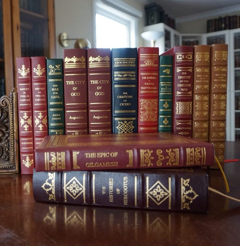 Puede incluir: Una colecci&oacute;n de libros encuadernados en cuero con letras doradas y dise&ntilde;os decorativos. Los libros est&aacute;n dispuestos en una fila sobre una superficie de madera. Los t&iacute;tulos incluyen "La Ciudad de Dios", "Las Oraciones de Cicer&oacute;n", "La Guerra del Peloponeso" y "La Epopeya de Gilgamesh".
