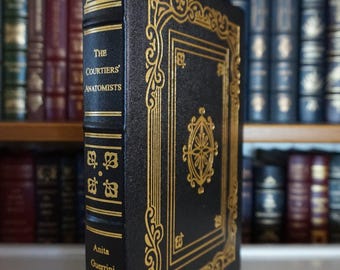 May include: A hardcover book titled "The Courtiers' Anatomists" by Anita Guerrini. The book has a black cover with gold decorative accents and text. The book is standing upright with a yellow bookmark.