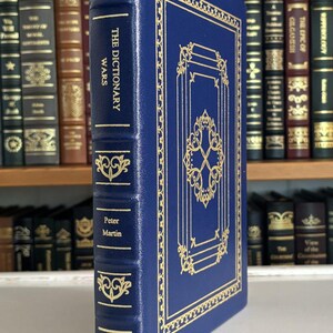 Puede incluir: Un libro de tapa dura azul titulado "The Dictionary Wars" de Peter Martin. El libro tiene detalles decorativos dorados y letras doradas. Se ve una cinta dorada como marcapáginas. Otros libros están en un estante en el fondo.