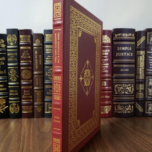 Pode incluir: Um livro de capa dura bordô intitulado "Teddy Roosevelt & The Marquette Libel Trial" de Mikel Classen. A capa apresenta detalhes em dourado e uma borda decorativa. Outros livros são visíveis ao fundo.