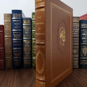 May include: A brown leather-bound book titled "The Aphorisms of Hippocrates" with gold lettering and a gold-leaf design. The book is part of a series called "The Classics of Medicine Library".