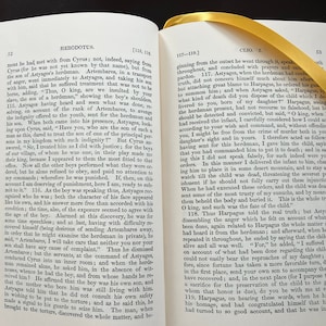 May include: A close-up of a page from a book titled "Herodotus" with text about the story of Cyrus, the son of Astyages, and his treatment by the son of a herdsman. The text is in black and white.