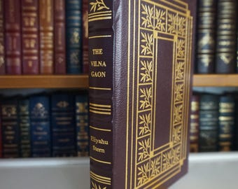 May include: A hardcover book titled "The Vilna Gaon" by Eliyahu Stern. The book has a dark burgundy cover with gold decorative accents and text. A yellow bookmark is inserted. The book is displayed in front of a bookshelf filled with other books.