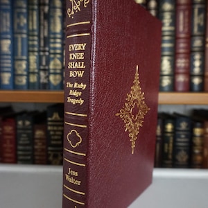 Può includere: Un libro con copertina rigida in pelle bordeaux. Il titolo "EVERY KNEE SHALL BOW" e l'autore "Jess Walter" sono in lettere dorate. Un disegno decorativo dorato è sulla copertina. Il libro fa parte di una collezione.