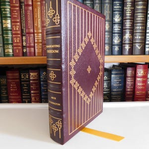 Könnte beinhalten: Ein weinrotes Hardcover-Buch mit dem Titel "Broadcasting Freedom" von Arch Eddington. Das Buch hat goldene Verzierungen und ein goldenes Lesezeichen. Weitere Bücher sind im Hintergrund zu sehen.