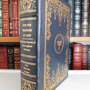 Puede incluir: Un libro de tapa dura de color azul oscuro con letras doradas y detalles decorativos. El t&iacute;tulo es "Por la libertad de Sion: La gran revuelta de los jud&iacute;os contra los romanos" de Guy MacLean Rogers. El libro tiene un marcap&aacute;ginas dorado.