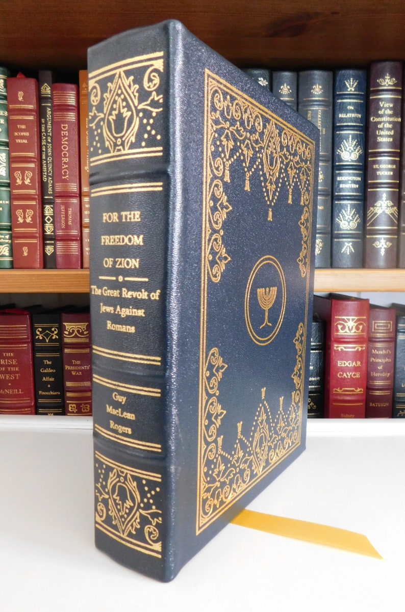 Puede incluir: Un libro de tapa dura de color azul oscuro con letras doradas y detalles decorativos. El t&iacute;tulo es "Por la libertad de Sion: La gran revuelta de los jud&iacute;os contra los romanos" de Guy MacLean Rogers. El libro tiene un marcap&aacute;ginas dorado.