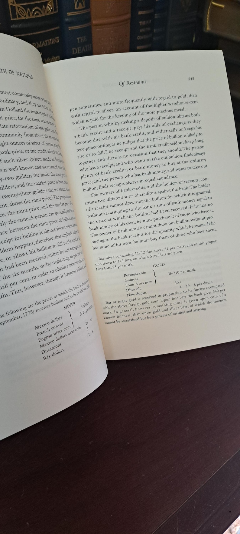 K&ouml;nnte beinhalten: Aufgeschlagenes Buch mit Text &uuml;ber Wirtschaft, einschlie&szlig;lich des Titels &bdquo;Of Restraints&ldquo; und Tabellen mit Silber- und Goldpreisen. Die Seiten sind vergilbt, das Buch ist blau gebunden.