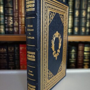 Könnte beinhalten: Ein dunkelblaues Hardcover-Buch mit goldenen Akzenten und dem Titel "Hulk Hogan v. Gawker" von Ryan Holiday. Das Buch befindet sich in einem Bücherregal mit anderen Büchern.