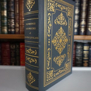 May include: A hardcover book titled "The Writings and Speeches of Grover Cleveland." The book has a dark blue cover with gold decorative floral patterns and text. The spine also features gold accents. The background shows a bookshelf filled with other books.