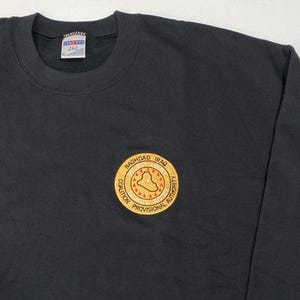 Könnte beinhalten: Schwarzes Sweatshirt mit Rundhalsausschnitt und einem gold-roten Aufnäher mit der Aufschrift "BAGHDAD IRAQ COALITION PROVISIONAL AUTHORITY". Das Sweatshirt hat ein Etikett mit der Aufschrift "JERZEES XL 262".