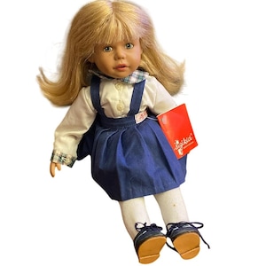 Puede incluir: Muñeca Sigikid con cabello rubio, vestida con blusa blanca, vestido peto azul, calcetines blancos y zapatos azules. La muñeca tiene una cara realista y sostiene una etiqueta roja.