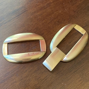 May include: Two gold-toned belt buckles. One is oval with the text "ELLEN TRACY" and the other is a similar shape with a rectangular tab. The buckles are on a brown surface.