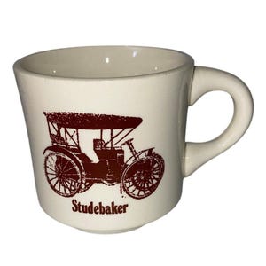 May include: A cream-coloured ceramic mug featuring a dark brown graphic illustration of a vintage Studebaker automobile on the front. The word Studebaker is printed in a dark brown serif font directly below the car illustration on the mug.