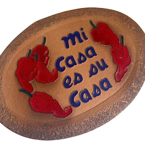 May include: An oval-shaped terracotta plaque with a textured brown surface. The plaque features the blue text "mi casa es su casa" in the center, surrounded by three glossy red chili pepper illustrations with green stems.
