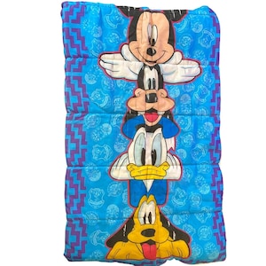 Puede incluir: Un saco de dormir acolchado rectangular en azul brillante que presenta una pila vertical de cuatro personajes de Disney: Mickey Mouse, Goofy, el Pato Donald y Pluto. El fondo tiene un patrón repetitivo de líneas en zigzag moradas y logotipos de Mickey Mouse.
