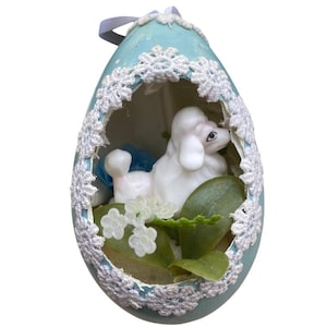 May include: A light blue, egg-shaped ornament with a white lace trim. Inside, a white poodle figurine rests among green leaves and white flowers. A blue ribbon is attached at the top for hanging. The ornament is a decorative item.