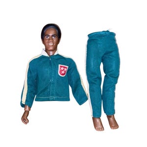 Op de afbeelding: Een pop met een donkere huid en zwart haar, gekleed in een blauwgroen jasje met witte zijstrepen en een rood schildembleem met drie witte sterren op de borst, gecombineerd met een bijpassende blauwgroene broek met witte zijstrepen.