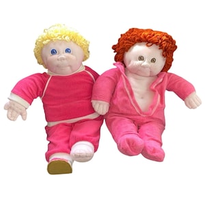 May include: Two vintage Cabbage Patch Kids dolls. One doll has blonde curly hair and is wearing a pink sweatsuit with white trim. The other doll has red curly hair and a pink jumpsuit. Both dolls have blue eyes and rosy cheeks.