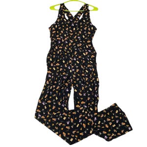 Könnte beinhalten: Schwarzer Jumpsuit mit gekreuztem Rücken und weiten Beinen. Der Jumpsuit ist mit einem Muster aus kleinen Teekannen, Blumen und anderen floralen Elementen in Orange, Gelb und Lila bedeckt. Er hängt an einem grünen Kleiderbügel.