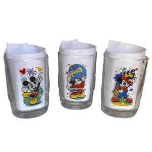 Puede incluir: Tres vasos de cristal transparente, cada uno con un inserto de papel blanco. El primer vaso muestra a Mickey Mouse señalando hacia arriba con fuegos artificiales. El segundo muestra a Mickey como mago con una varita. El tercero muestra a Mickey con una claqueta.