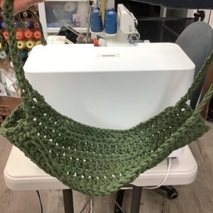 May include: An olive green crochet hammock with braided rope handles. The hammock is suspended, displaying its open weave and robust construction. The background includes sewing equipment, suggesting a craft or home decor item.