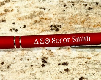 Pode incluir: Uma caneta vermelha com detalhes prateados e ponta preta. A caneta tem o texto branco "ΔΣΘ Soror Smith". Está sobre uma superfície salpicada de cor castanho claro.