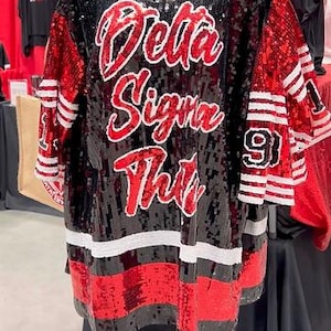 May include: Black sequined jersey with red accents and the words "Delta Sigma Theta" in red script. The jersey has white stripes on the sleeves and bottom, with the number 91 on the sleeve.