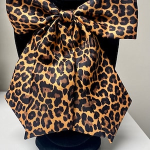 May include: A large, brown and black leopard print bow tie made of satin fabric. The bow tie is tied and displayed on a black mannequin.
