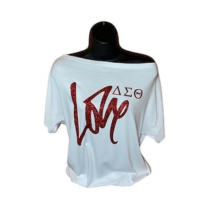 May include: White off-the-shoulder t-shirt with a red glitter "Love" graphic and Greek letters. The short-sleeved top is displayed on a black mannequin. The shirt is made of a soft material.