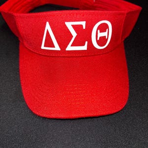 May include: A red visor with white Greek letters. The visor has a curved brim and a band to fit the head. The letters are Delta, Sigma, and Theta.