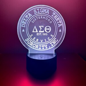 Pode incluir: Uma luz noturna de acr&iacute;lico branco com um design circular que apresenta o texto "Delta Sigma Theta", "Sisterhood", "Service", "Sorority, Inc." e "Est. 1913". O design tamb&eacute;m inclui as letras gregas "&Delta;&Sigma;&Theta;". A luz noturna tem uma base preta com um interruptor de ligar/desligar.