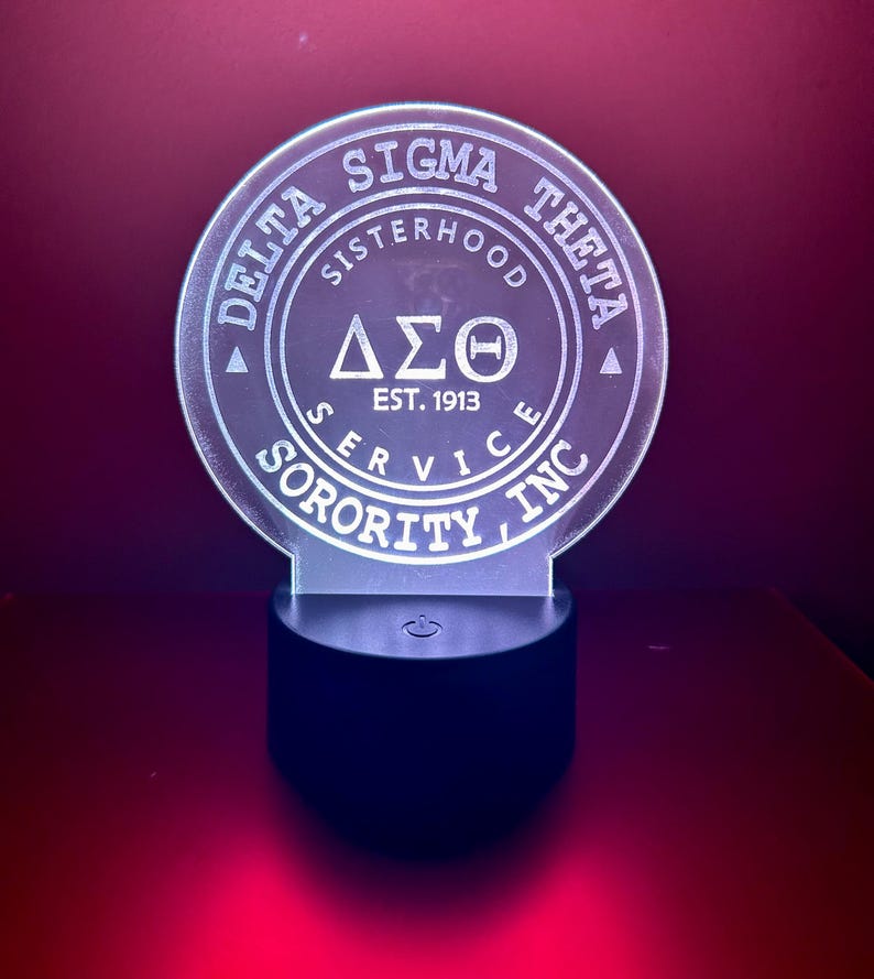 Pode incluir: Uma luz noturna de acr&iacute;lico branco com um design circular que apresenta o texto "Delta Sigma Theta", "Sisterhood", "Service", "Sorority, Inc." e "Est. 1913". O design tamb&eacute;m inclui as letras gregas "&Delta;&Sigma;&Theta;". A luz noturna tem uma base preta com um interruptor de ligar/desligar.