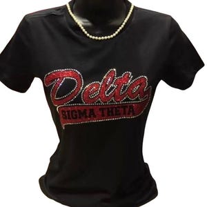May include: Black t-shirt with the words "Delta Sigma Theta" in red and rhinestone lettering. A pearl necklace is draped around the mannequin's neck. The shirt is short-sleeved.