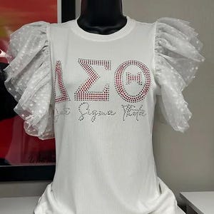 May include: White t-shirt with a rhinestone design of the Greek letters Delta Sigma Theta and the words "Delta Sigma Theta" underneath. The shirt has white tulle ruffle sleeves with polka dots.