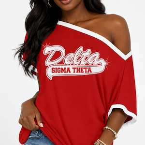 May include: A red off-the-shoulder t-shirt with white trim on the neckline and sleeves. The front features the text "Delta" in a white script font and "SIGMA THETA" in a white block font, both outlined with a sparkling rhinestone effect.